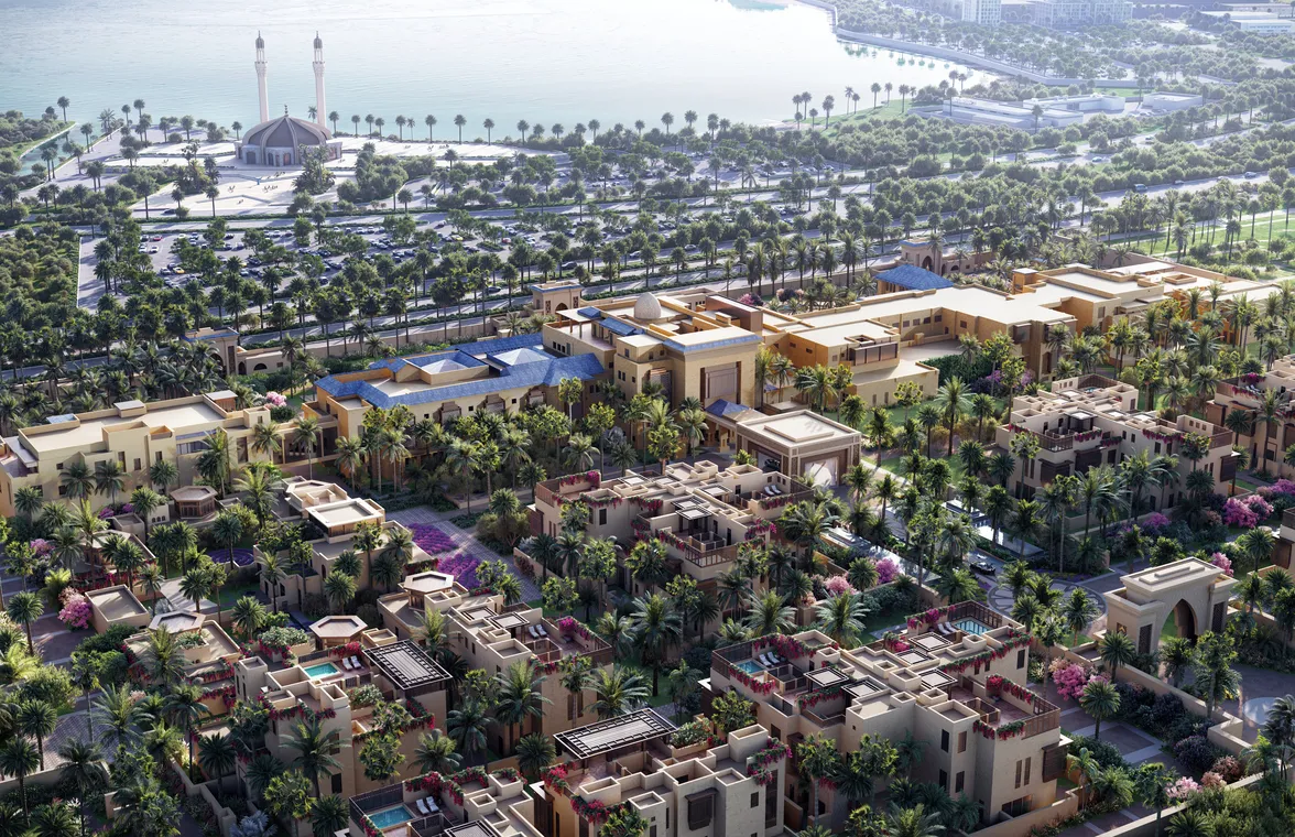 Traditional desert architecture with earth-toned villas and dense palm landscaping near a waterfront mosque and coastal road.